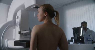 Yetişkin kadın, hastane radyoloji odasında üstsüz mammografi taraması yapıyor. Radyolog bilgisayarla mammogram makinesi kuruyor. Göğüs kanseri önleme. Modern parlak klinik.
