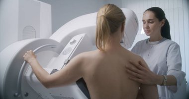 Hastane radyoloji odası. Mammografi taraması sırasında beyaz kadın ayakta duruyor. Kadın doktor dijital mammografiyi hastaya göre ayarlıyor, bilgisayar kullanıyor. Göğüs kanseri önleme. Modern klinik.