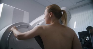 Yetişkin kadın, klinik radyoloji odasında mammografi çektirirken üstsüz duruyor. Erkek doktor bilgisayarla mamografi makinesi kuruyor. Göğüs kanseri önleme. Modern, parlak hastane.