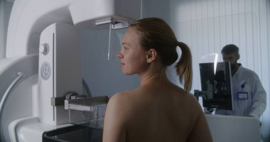 Yetişkin bir kadın klinik radyoloji odasında üstsüz mammografi taraması yaptırıyor. Erkek doktor mamogram makinesini bilgisayar kullanarak ayarlar. Göğüs kanseri önleme. Modern, parlak hastane.