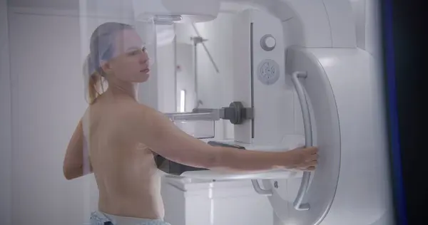 Caucasian adult woman stands topless in hospital radiology room. Female patient undergoing mammography screening procedure using mammogram machine. Breast cancer prevention. Modern clinic equipment.