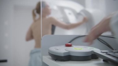 Yetişkin kadın, hastane radyoloji odasında mammografi taraması sırasında üstsüz duruyor. Radyolog kontrol panelindeki düğmeye basarak dijital mammografı çalıştırıyor. Modern klinik.