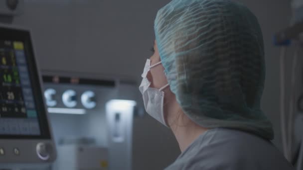 戴着医用帽和面罩的女医生用现代设备在手术室里看着心电图监测器 成年护士在手术期间与外科医生交谈 在诊所工作的医务人员 — 图库视频影像