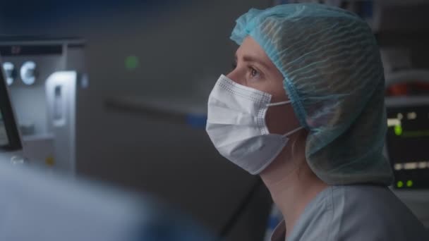 戴着医疗帽和口罩的女性成年医生坐在现代手术室 与外科医生交谈 护士检查病人在困难手术期间的心电图监测和检查情况 — 图库视频影像