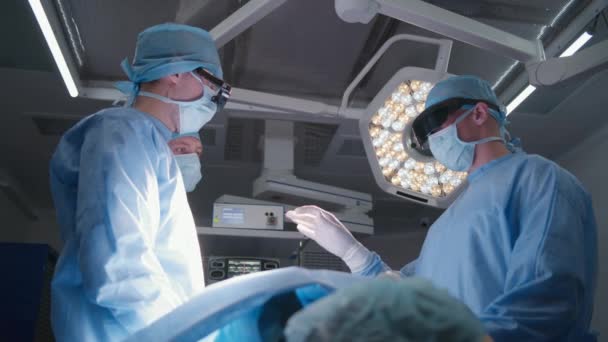 保護眼鏡の外科医チームは 患者とのテーブルの近くに立って 外科手術のプロセスを議論しています 看護師 救急隊員が手術に備えています 医療施設のオペレーティングルーム — ストック動画