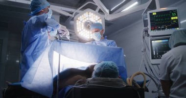 Hemşire hastanın durumunu elektrokardiyografi monitöründen kontrol ediyor. Yetişkin adam masada anestezi altında yatıyor. Profesyonel cerrahlar laparoskopi yapar ve monitörlere bakarlar. Sıhhiye ameliyathanesi.