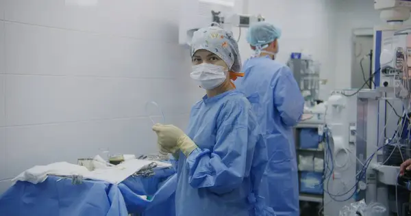 Professional male surgeon and nurses stand in operating room after long operation. Female medics clean workspace after surgery. Doctor takes off gloves. Medical staff at work in modern clinic.