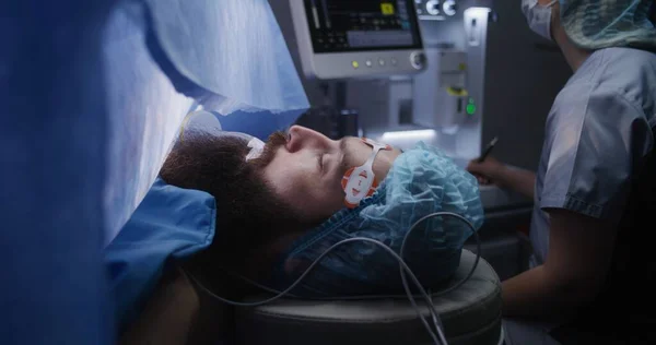围手术期病人在麻醉下躺在手术台上用呼吸管进行麻醉 护士看着监视器 写下心电图结果 手术室工作的医务人员 — 图库照片