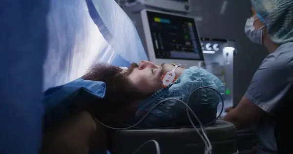 手術中に呼吸管で麻酔の下で外科テーブルに横たわっている患者の近く 看護師はモニターを見て 電気泳動の結果を書いています オペレーティングルームで働く医療スタッフ — ストック写真