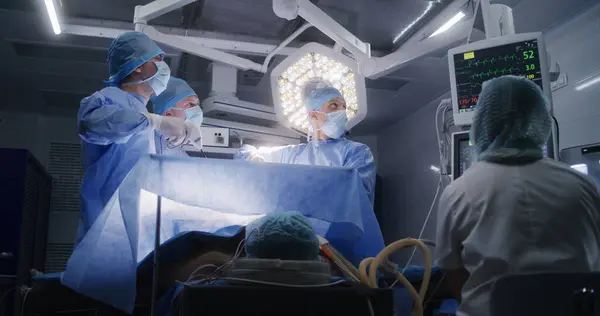 看護師は 電気泳動モニターの患者の状態をチェックします 大人の男はテーブルの上の麻酔の下に横たわっています プロの外科医は腹腔鏡検査を行い モニターを見ています メディックオペレーティングルーム — ストック写真