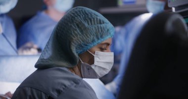 Kadın doktor modern ameliyathanede oturuyor ve hastanın durumunu elektrokardiyografi monitöründen kontrol ediyor. Cerrahi kıyafetli profesyonel erkek cerrahlar laparoskopi aletleri kullanarak ameliyat yaparlar..