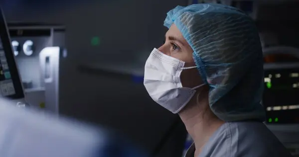 Female adult medic in medical hat and mask sits in modern operating room and talks with surgeons. Nurse looks at electrocardiography monitor and checks condition of patient during difficult surgery.