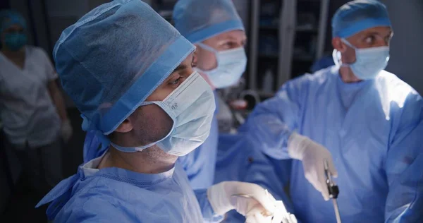Close up of male surgeon in surgical suit performing laparoscopy operation with colleagues. Professional medics operate patient and look at monitors in modern operating room. Medical staff at work.