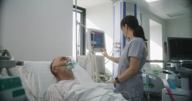 Klinikte modern ekipmanlarla acil servis. Oksijen maskeli yaşlı adam akciğer havalandırması sırasında yatakta yatıyor. Dost canlısı hemşire, yaşam destek makinesini eski hastaya göre ayarlıyor. Yoğun Bakım Bölümü.