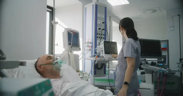 Emergency room with modern equipment in clinic. Senior man in oxygen mask lies in bed during lung ventilation. Friendly nurse adjusts life support machine for old patient. Intensive care department.