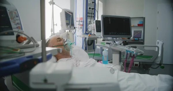 急救室 诊所配备现代化设备 戴氧气面罩的老年人在呼吸空气时躺在床上 友好的护士为老年病人调整生命保障机 重症监护科 — 图库照片