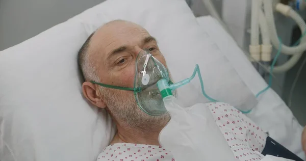 Nurse takes care of sick senior patient. Elderly man in oxygen mask lies in bed during artificial lung ventilation. Emergency room in modern hospital. Intensive care coronavirus department in clinic.