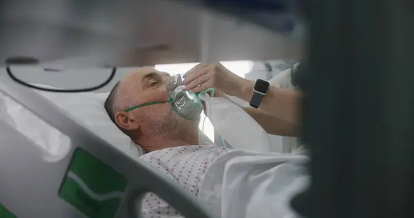 Old patient in oxygen mask lies in bed during lung ventilation. Nurse takes care of elderly man, uses monitor. Emergency room with modern equipment in hospital. Intensive care coronavirus department.
