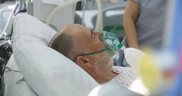 Friendly nurse talks and takes care of sick elderly patient wearing oxygen mask, uses hospital monitor. Emergency room with modern equipment in clinic. Intensive care department in medical facility.