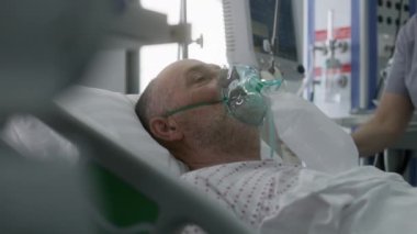 Oksijen maskeli yaşlı adam yapay akciğer havalandırması sırasında yatakta yatıyor. Hemşire hastayla ilgilenir. Modern klinikteki acil servis. Tıbbi tesiste yoğun bakım Coronavirus Bölümü.