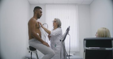 Kadın kardiyolog, doktor kardiyovasküler hastalığı olan hastaya holter sensör takıyor. Kardiyoloji odasındaki işlevsel rehabilitasyon sırasında Afrikalı Amerikalı bir adam. Modern hastanede elektrokardiyografi