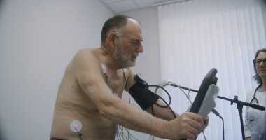 Holter sensörlü yaşlı bir adam kardiyoloji odasında yörünge pedalları çeviriyor. Kadın kardiyolog kardiyovasküler hastalığı olan hastayı teşhis ediyor. Modern hastanede işlevsel rehabilitasyon.