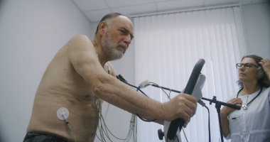 Holter sensörlü yaşlı bir adam kardiyoloji odasında yörünge pedalları çeviriyor. Kadın kardiyolog kardiyovasküler hastalığı olan hastayı teşhis ediyor. Modern hastanede işlevsel rehabilitasyon.