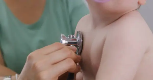 Mom holds pretty baby with pacifier on her hands. Pediatrician uses stethoscope to listen to heartbeat and lungs of handsome little kid. Doctor does checkup in modern hospital. Close up. Slow motion.