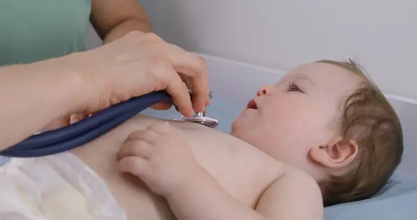 Cute little child lies on changing table and coughs. Pediatrician uses stethoscope to listen heartbeat and lungs of handsome cute baby. Physician does checkup in medical center. Close up. Slow motion.