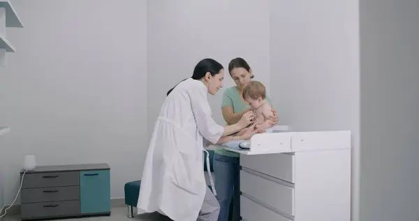 Mother holds her cute baby in diaper and puts him on changing table. Pediatrician uses stethoscope to listen to heartbeat and lungs of little child. Physician does checkup of kid in modern hospital.