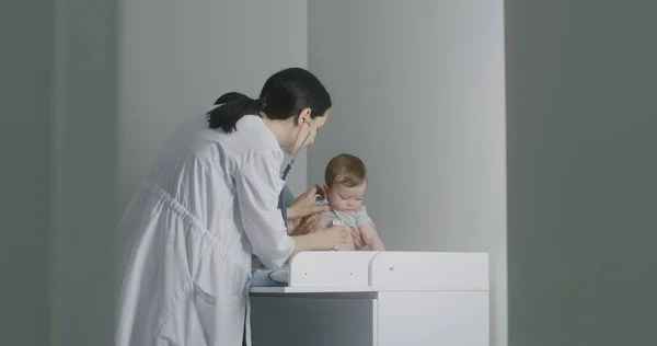Friendly female pediatrician gives toy to handsome baby sitting on changing table. Physician uses stethoscope to listen to heartbeat and lungs of cute little child. Doctor does checkup in hospital.