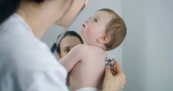 Pediatrician applies stethoscope to back of handsome little child to listen heartbeat and lungs. Mother holds on her hands cute baby on doctor checkup. Physician works in modern hospital. Close up.