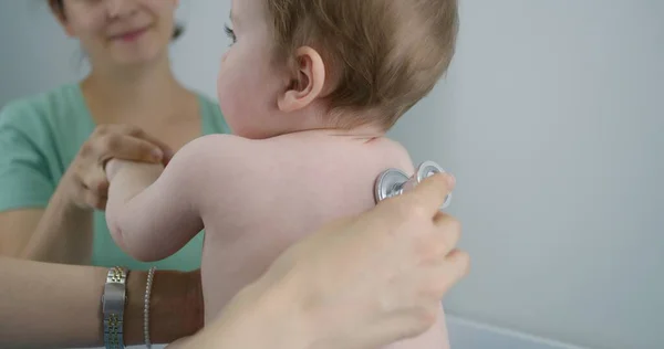 Pediatrician applies stethoscope to back of handsome little child to listen heartbeat and lungs. Mother holds on her hands cute baby on doctor checkup. Physician works in modern hospital. Close up.