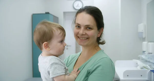 赤ちゃんを持つ大人の女性は 現代の明るい病院や診療室の真ん中に立っています 母親は子供を抱き カメラと笑顔を見つめている 小児科医との診察を受けた小さい子供の女性 ポートレイト — ストック写真