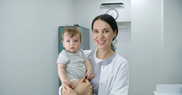 女性小児科医は 明るい病院の部屋の真ん中に立って カメラを見ています 大人の医師が赤ちゃんを抱えている 小さな男の子はカメラを見て 薬を握っています 近代クリニックで働く医療スタッフ — ストック写真