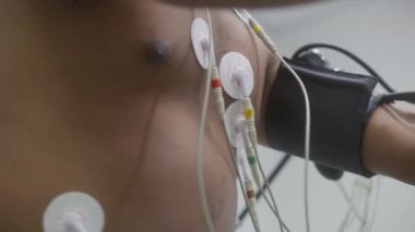 Tonometresi ve sensörleri olan Afrikalı Amerikalı bir adam elektrokardiyografi sırasında kardiyoloji odasında yörünge pedalları çeviriyor. Kardiyovasküler bozukluğu olan hasta. Modern klinikte işlevsel rehabilitasyon.