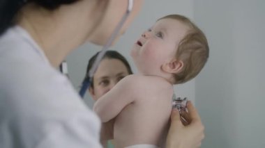 Pediyatrist, kalp atışlarını ve akciğerlerini dinlemek için küçük yakışıklı çocuğun sırtına steteskop takıyor. Anne ellerini tutuyor. Doktor muayenesinde tatlı bir bebek. Doktor modern hastanede çalışıyor. Kapat..