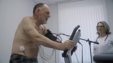 Holter sensörlü yaşlı bir adam işlevsel rehabilitasyon sırasında kardiyoloji odasında yörünge pedalları çeviriyor. Kadın doktor kalp rahatsızlığı olan hastaları muayene ediyor. Tıp merkezinde elektrokardiyografi.