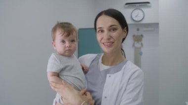 Yetişkin doktor bebekle birlikte modern hastane koğuşunun ortasında duruyor. Kadın çocuk doktoru kucağına alır, kameraya bakar ve gülümser. Klinikte, hastanede ya da tıp merkezinde çalışan tıbbi personel.