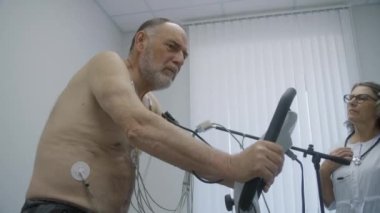 Holter sensörlü yaşlı bir adam kardiyoloji odasında yörünge pedalları çeviriyor. Kadın kardiyolog kardiyovasküler hastalığı olan hastayı teşhis ediyor. Modern hastanede işlevsel rehabilitasyon.
