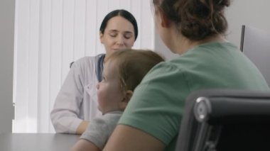 Bayan çocuk doktoru, küçük tatlı çocuğu olan bir hastaya hastanede danışmanlık yapıyor. Kadın doktor kontrolünde sandalyede oturuyor. Dost canlısı doktor modern tıp merkezinde çalışıyor. Kapatın. Arka plan.