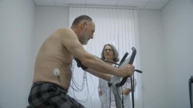 Kıdemli hasta, hastane odasında Holter monitörleri ve sensörleriyle yörüngeye oturuyor. Kadın kardiyolog ayakta duruyor, erkeğin durumunu kontrol ediyor. Hemşire bilgisayardan elektrokardiyogram sonuçlarını izliyor.