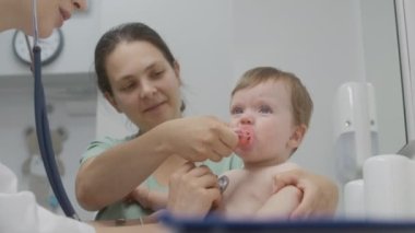 Küçük çocuk masasını değiştiriyor ve çocuk doktoruyla randevusunda ağlamaya başlıyor. Anne çocuğu kucağına alır ve emzik verir. Kadın doktor steteskop kullanıyor. Hastanedeki bebeğin durumunu kontrol ediyor..