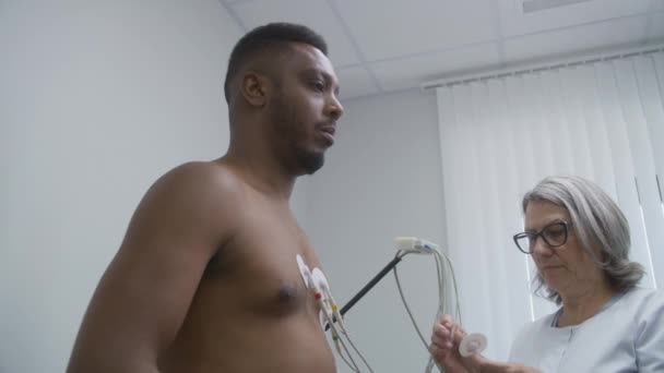 女性の心臓科医 医師は心血管疾患を患っている患者にホルターセンサーを置きます アフリカ系アメリカ人男性は 心臓病室で機能リハビリを行っている 現代病院における電気泳動 — ストック動画