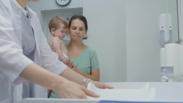 妈妈把孩子抱在手里 医生把秤放在更衣室的桌子上 女医生在现代儿科病房给孩子们称重量 与儿科医生预约的妇女与儿子 在医院工作的医务人员 — 图库视频影像