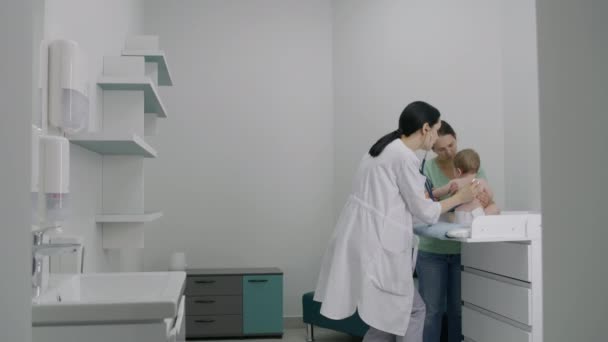 妈妈把她可爱的孩子抱在尿布里 把他放在更衣室的桌子上 儿科医生用听诊器倾听幼儿的心跳和肺 医生在现代医院给孩子们做检查 — 图库视频影像