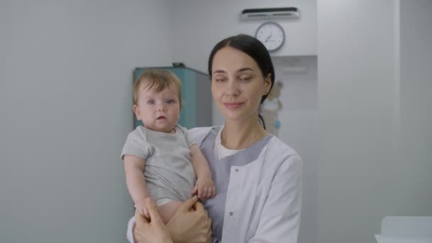 女儿科医生站在明亮的病房中央 看着镜头 成年医生抱着孩子 小男孩看着摄像机 牵着医生的手 在现代诊所工作的医务人员 — 图库视频影像