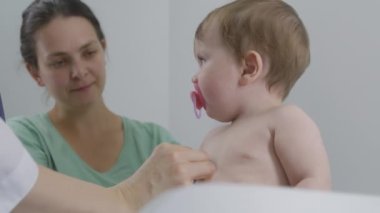 Anne, emziği olan yakışıklı bebeği masanın üzerinde otururken tutuyor. Pediyatrist kalp atışlarını ve küçük çocuğun ciğerlerini dinlemek için steteskop kullanıyor. Doktor klinikte check-up yapıyor. Kapatın. Yavaş çekim.