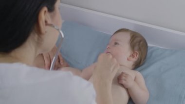 Annem, doktor kontrolünde, emzik taşıyan ve masasını değiştiren tatlı bir bebeğin elini tutuyor. Kadın çocuk doktoru, kalp atışlarını ve küçük tatlı çocuğun ciğerlerini dinlemek için steteskop kullanıyor. Kapatın. Yavaş çekim.