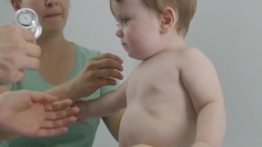 Anne bebeği masasının üstüne koyar ve ağzına emzik verir. Pediyatrist kalp atışlarını ve küçük çocuğun ciğerlerini dinlemek için steteskop kullanıyor. Doktor hastanede check-up yapıyor. Kapatın. Yavaş çekim.